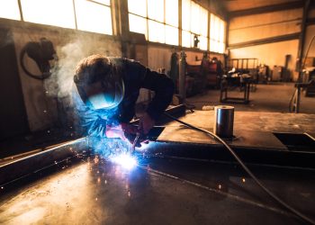 Professional welder in protective uniform and helmet welding metal part in workshop.